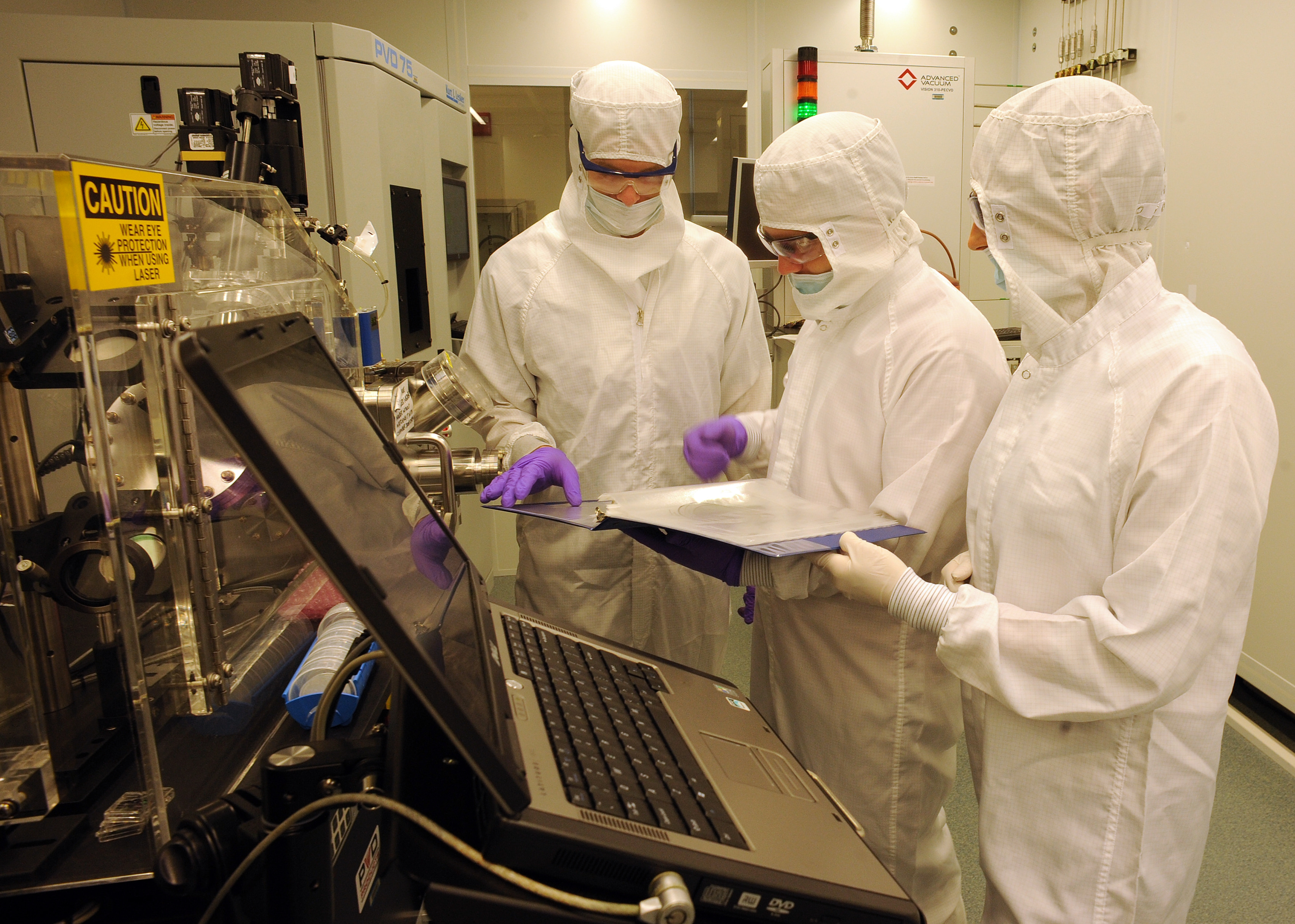 Three individuals in cleanroom suits examining a paper in a lab with equipment and a computer.