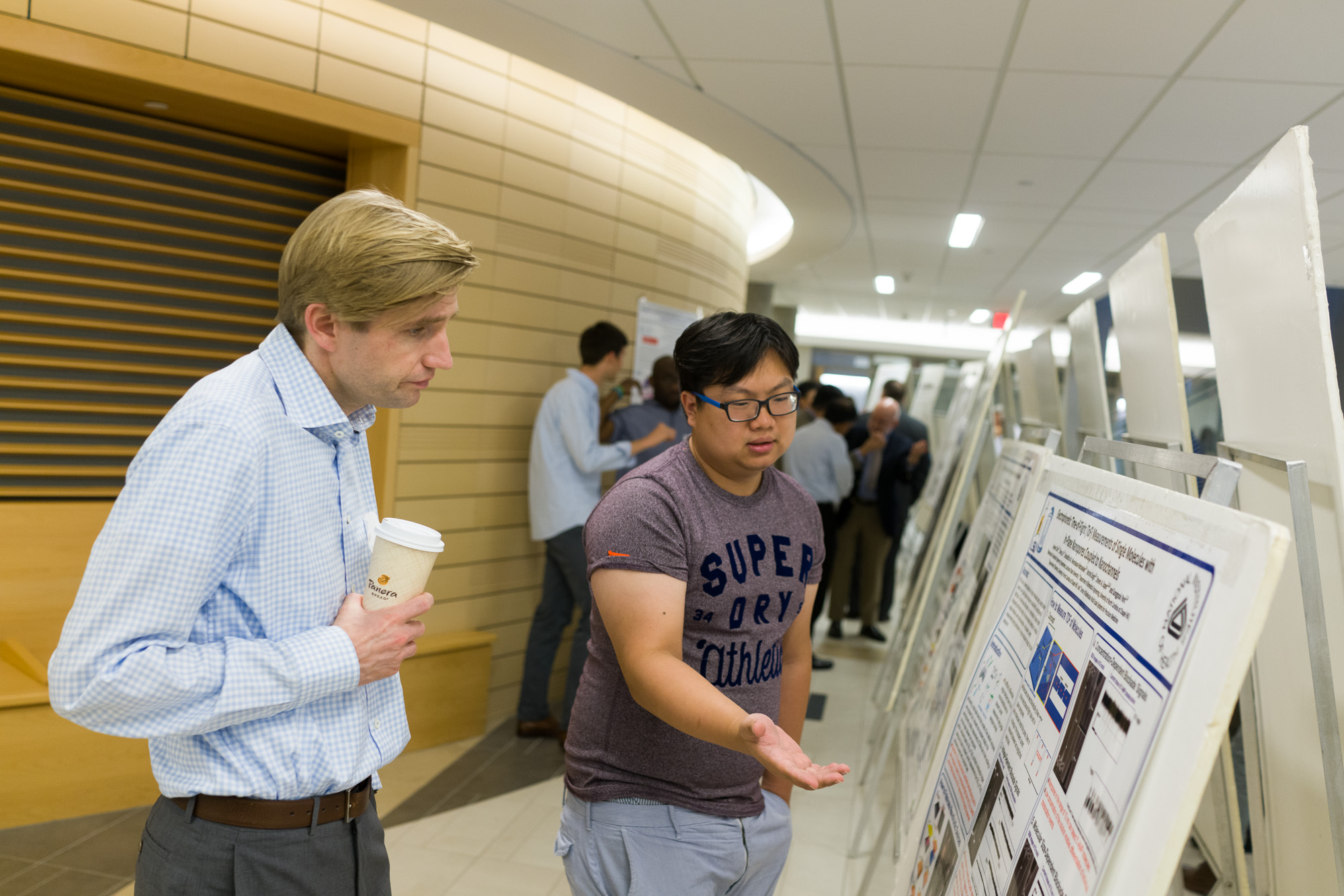 Two men discuss a scientific poster at a conference, with one holding a coffee cup and the other explaining with a hand gesture.