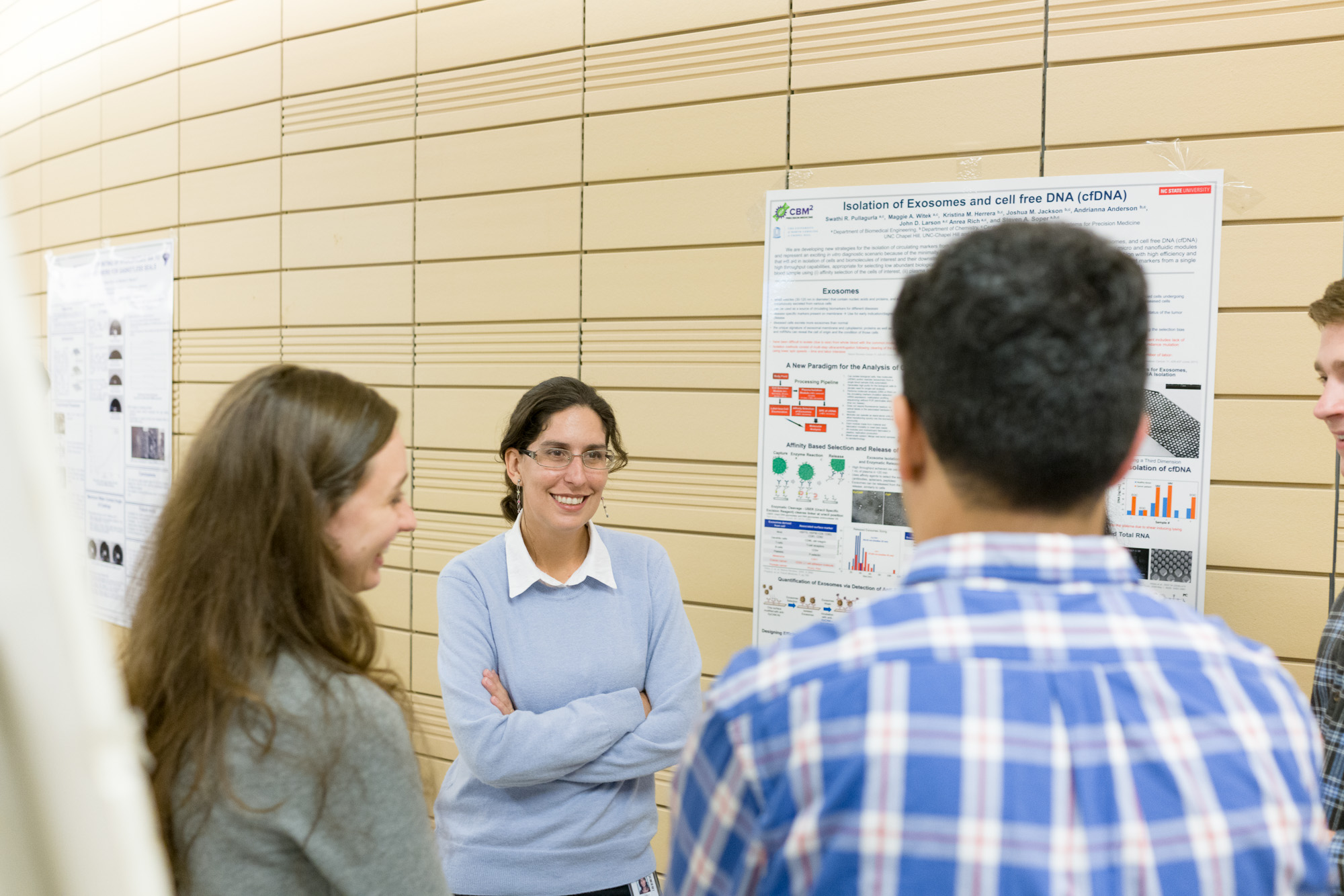 A group discusses a scientific poster on exosomes and cell-free DNA. A woman in a light blue sweater interacts with others in an academic setting.