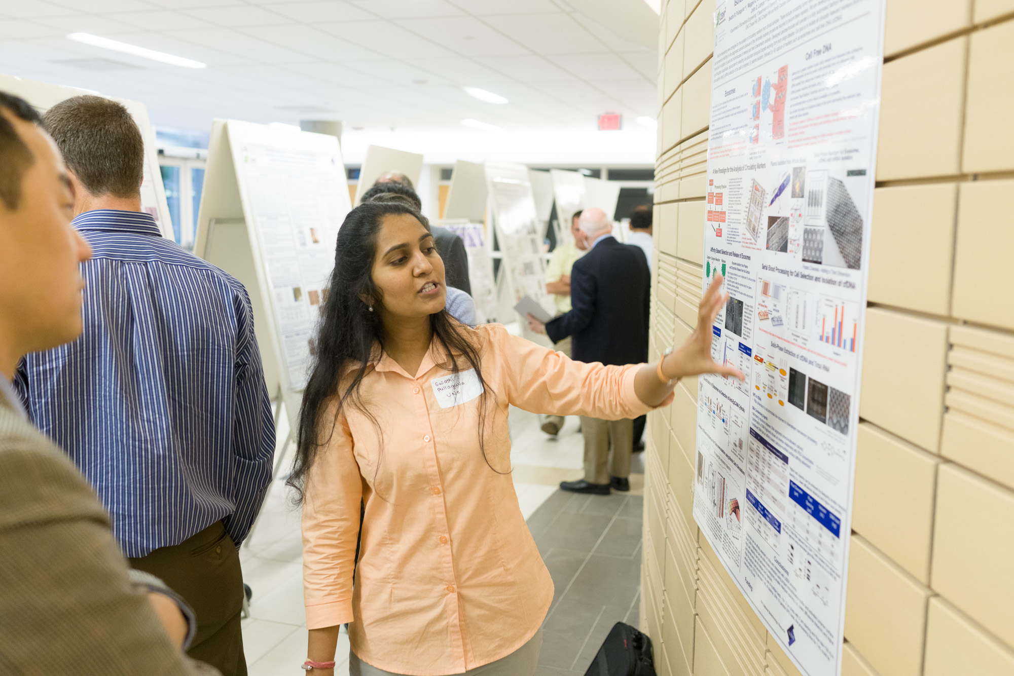 A woman is presenting a research poster at what appears to be an academic event, with other attendees and posters visible in the background.