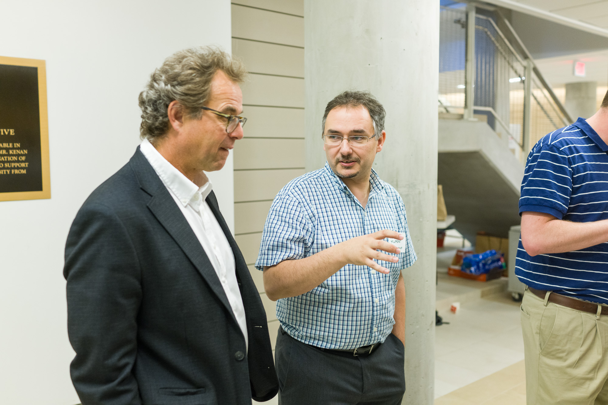 Two men in a modern building engage in a conversation; one wears glasses and a formal jacket, while the other holds a beverage and wears a striped shirt.