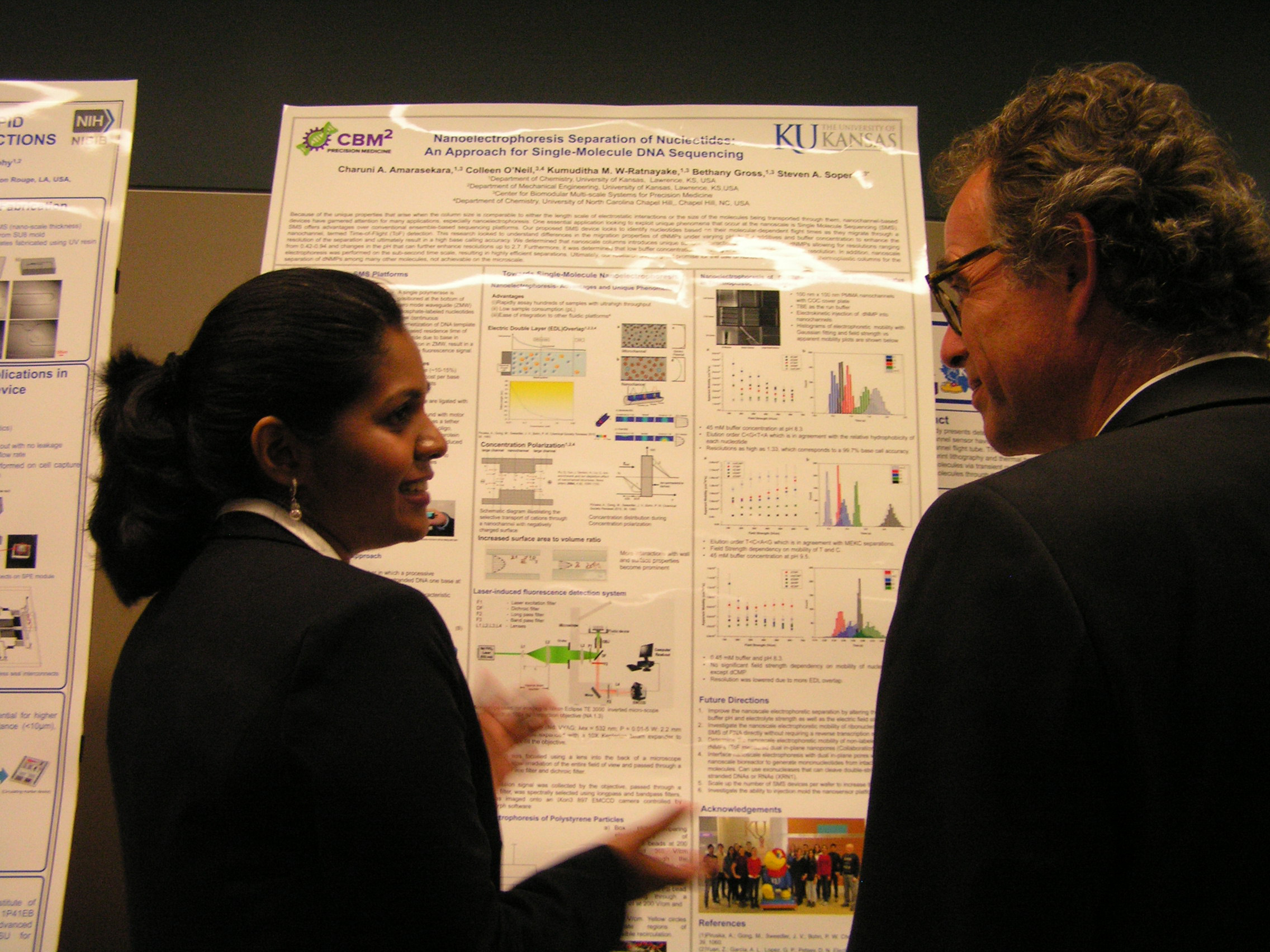 Two individuals in front of a research poster, one verbally explaining its contents.