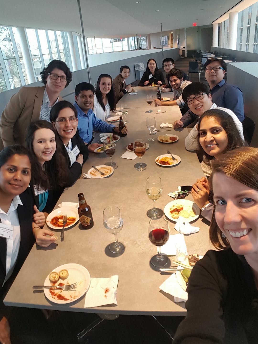 A group of people gathered around a meal, all smiling and participating in a group selfie taken on one individual's phone.