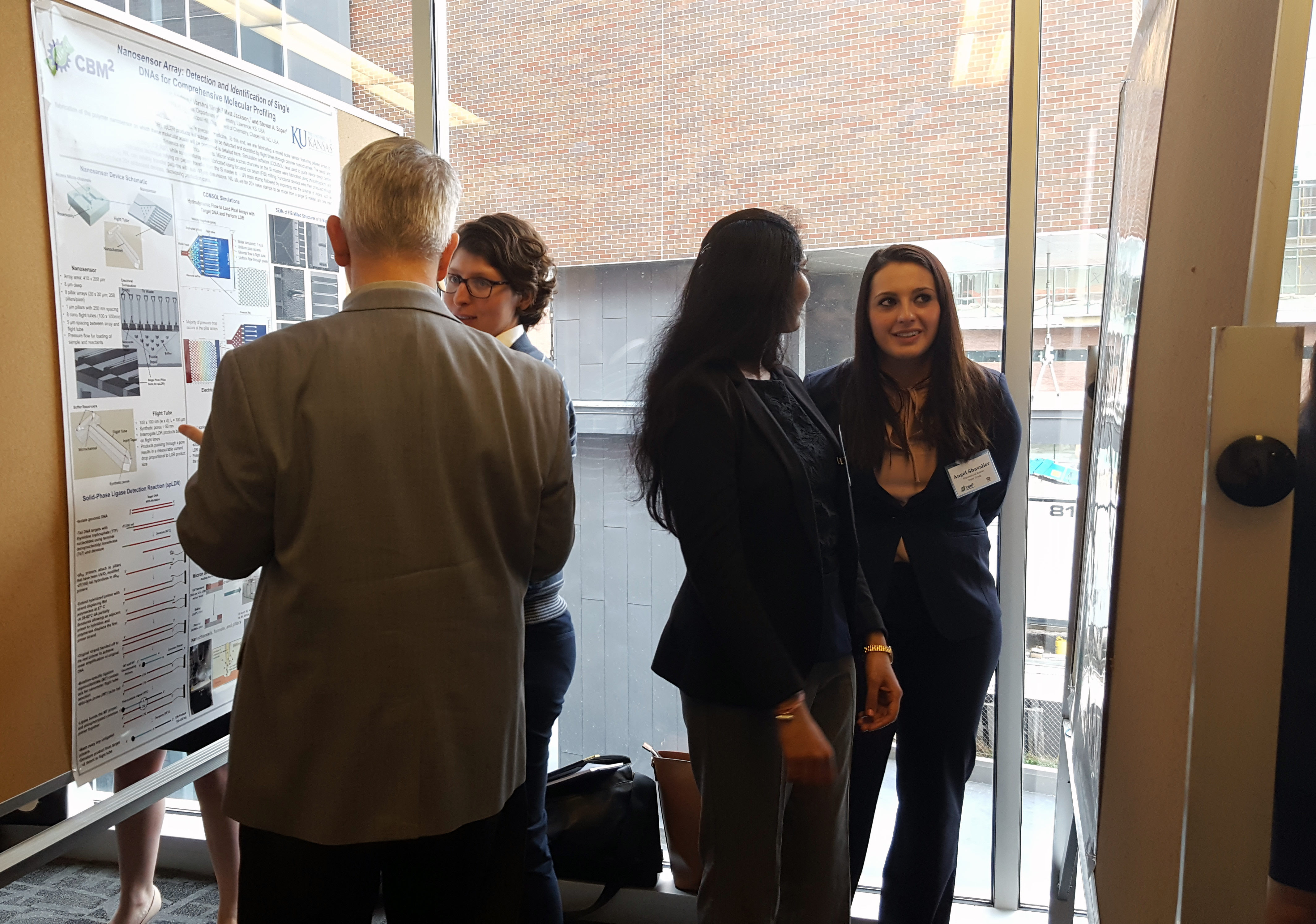 Four individuals split into two pairs, with each pair consisting of one person explaining the content on a research poster to an attentive listener.