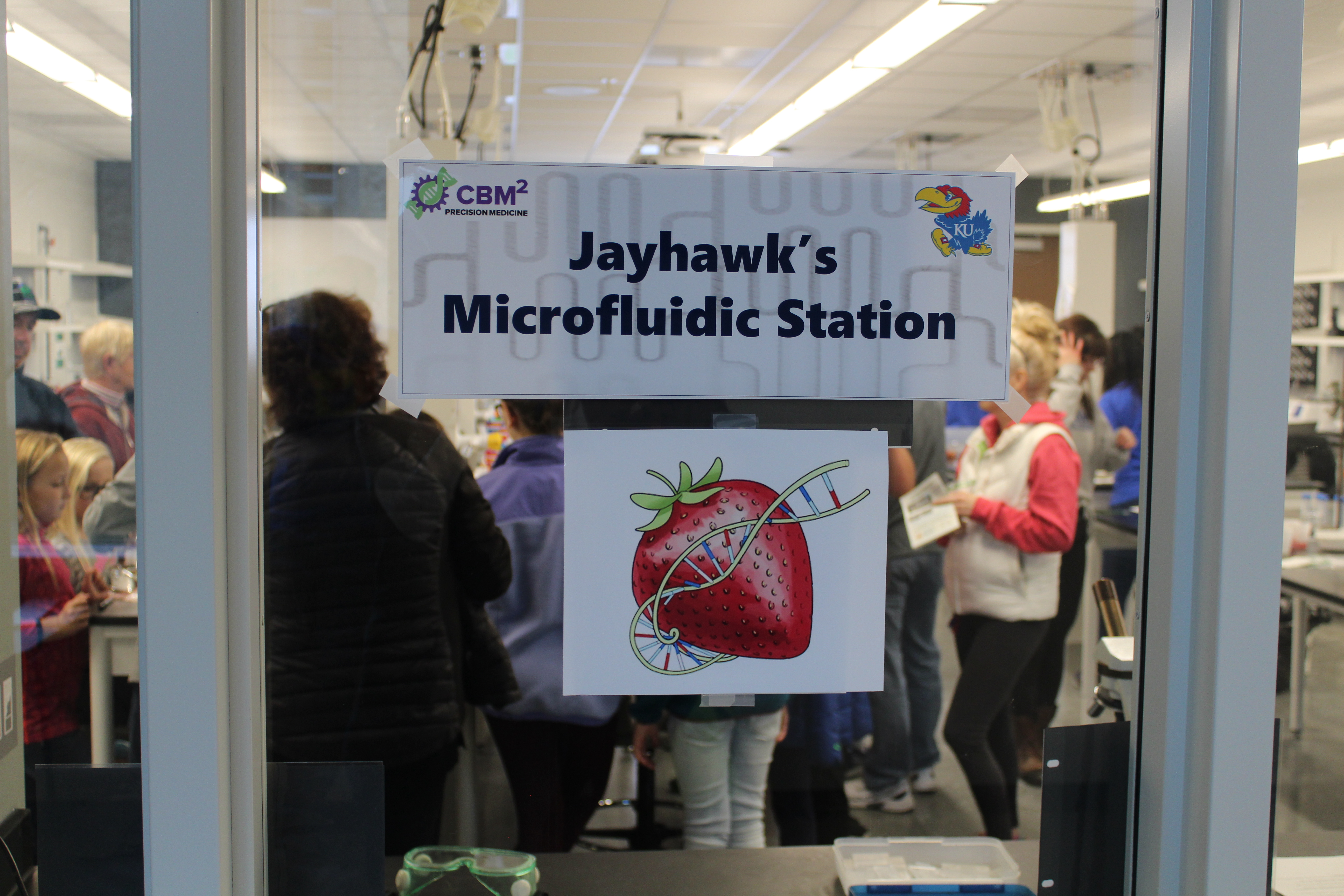 Sign reading 'Jayhawk's Microfluidic Station' with a strawberry encircled by a DNA strand.