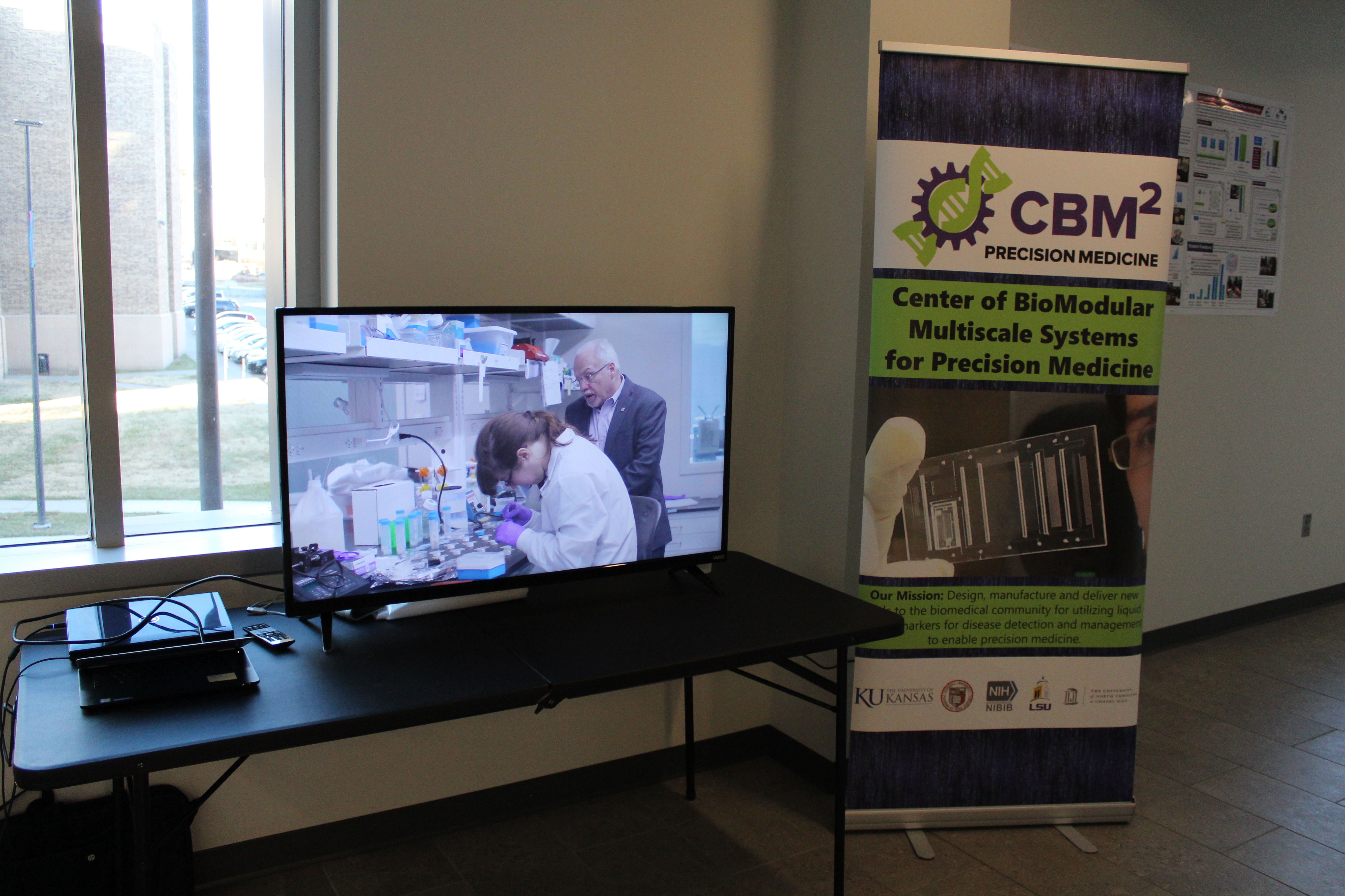 A television screen displaying a CBMM advertisement with a CBMM Chemistry Festival poster placed on a stand next to the table where the TV is located.