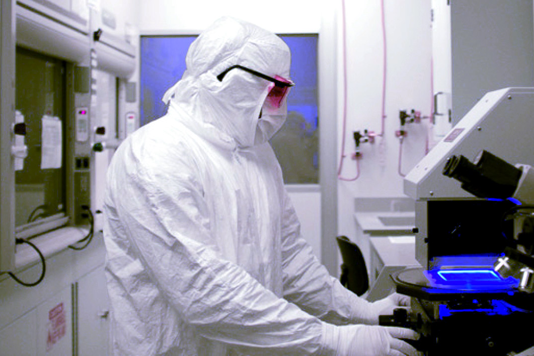 An individual in a cleanroom suit working with a microscope emitting a blue light in a lab setting.
