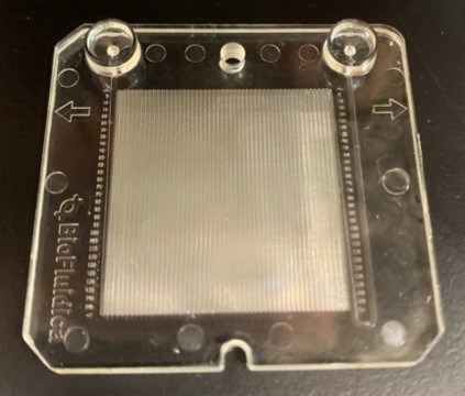 Clear plastic object with a mesh center and four corner holes on a dark surface.
