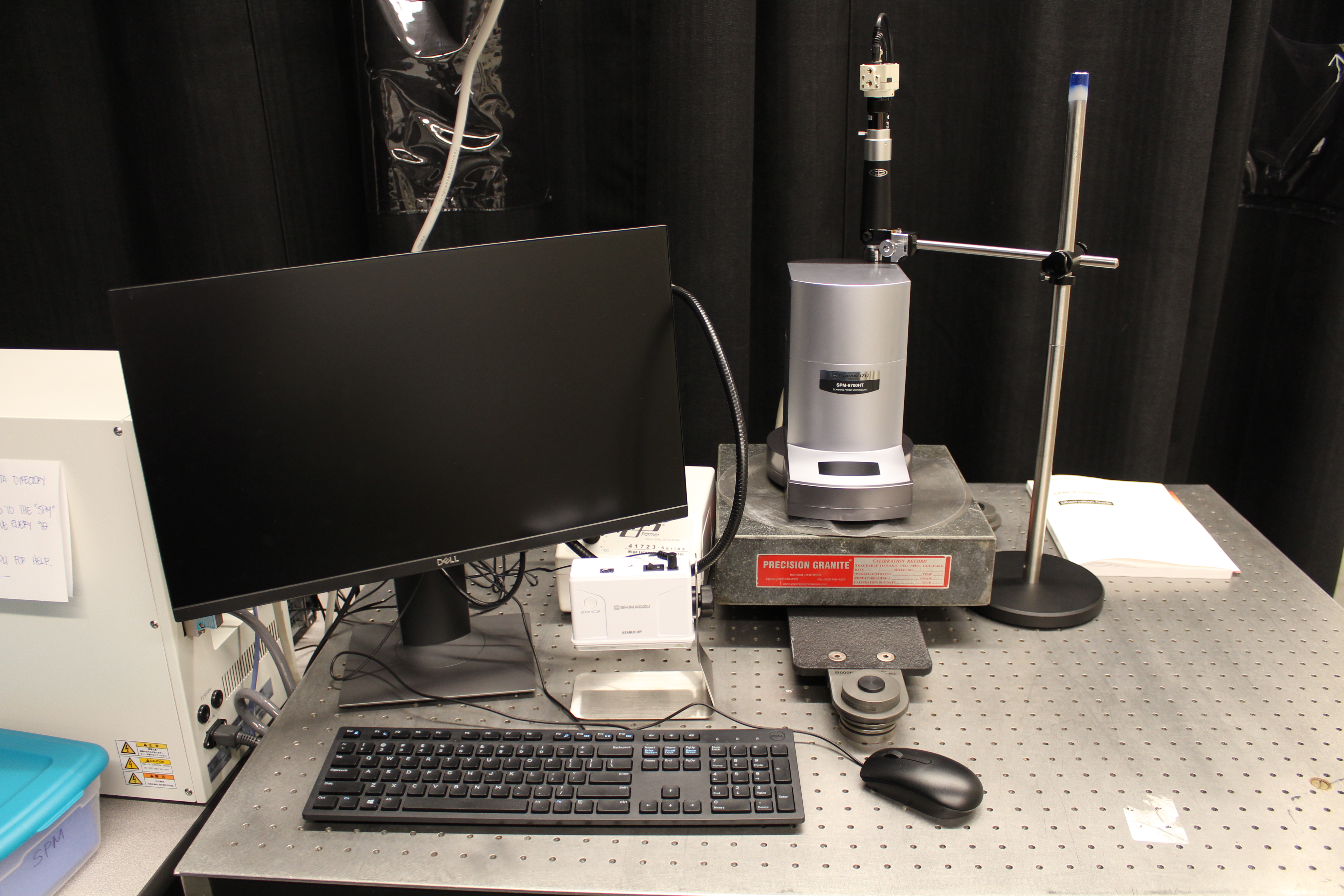 Lab setup with a monitor, measurement device on a stand, keyboard, mouse, and various tools on a perforated metal table.