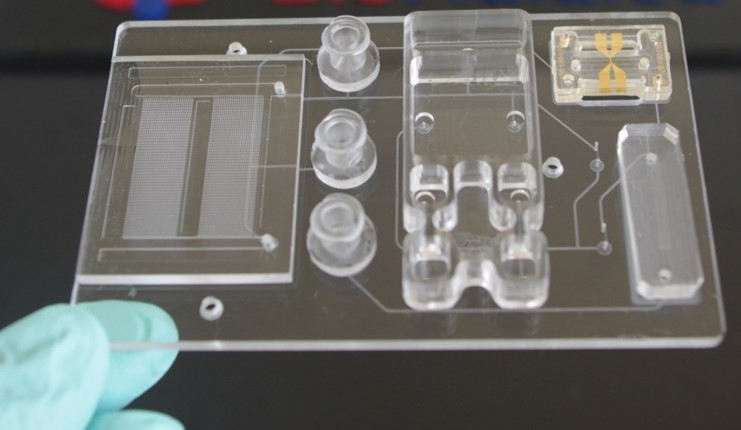 Transparent lab device with chambers and circuitry on a dark surface, with a gloved hand holding a pipette nearby.
