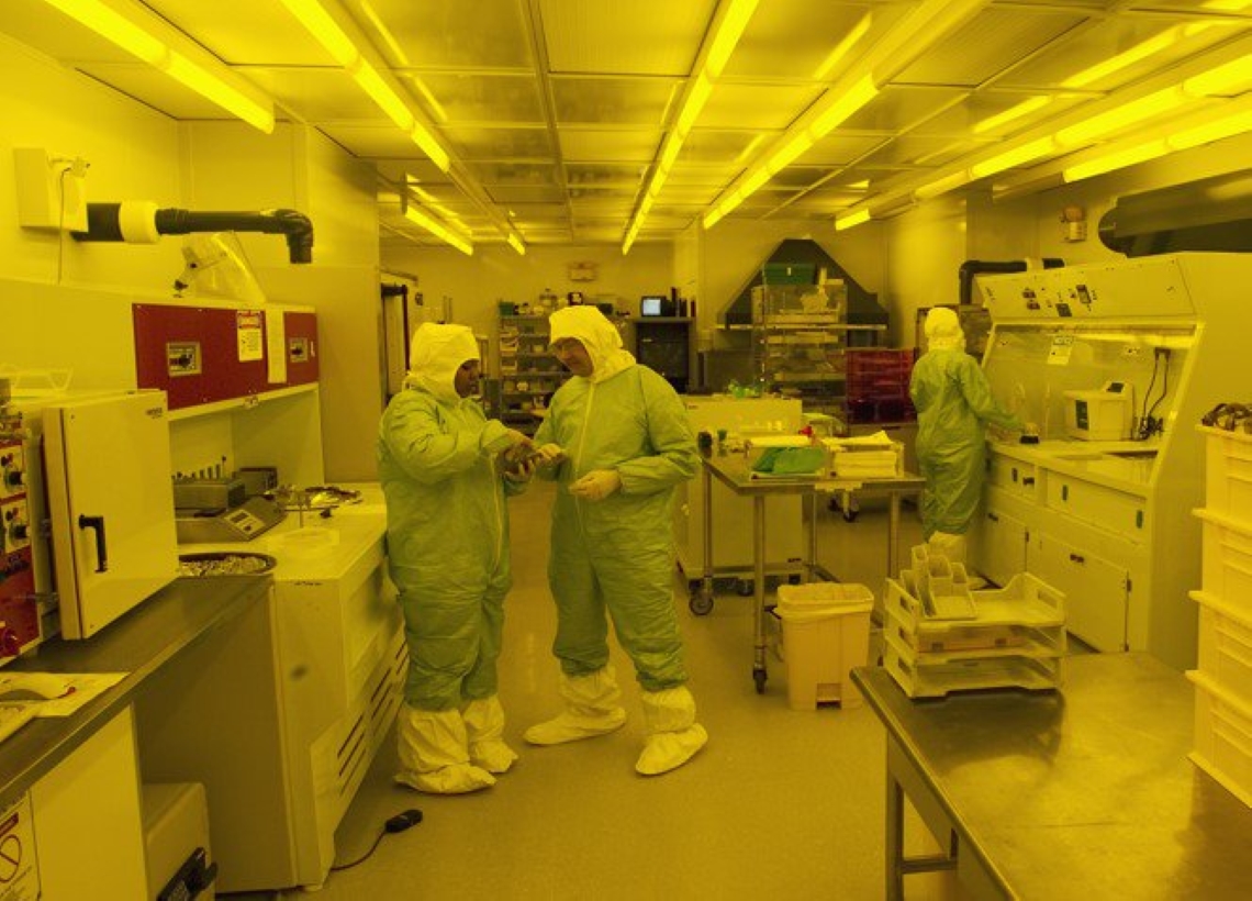 Three individuals in green cleanroom suits converse in a yellow-lit lab filled with equipment and workstations.