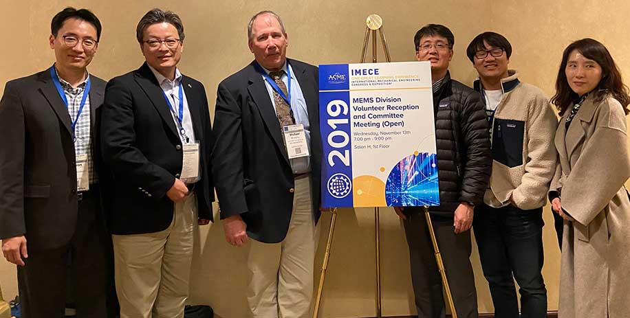 Group photo of five professionals at the ASME IMECE 2019 event, standing next to a sign for the MEMS Division Volunteer Reception and Committee Meeting.