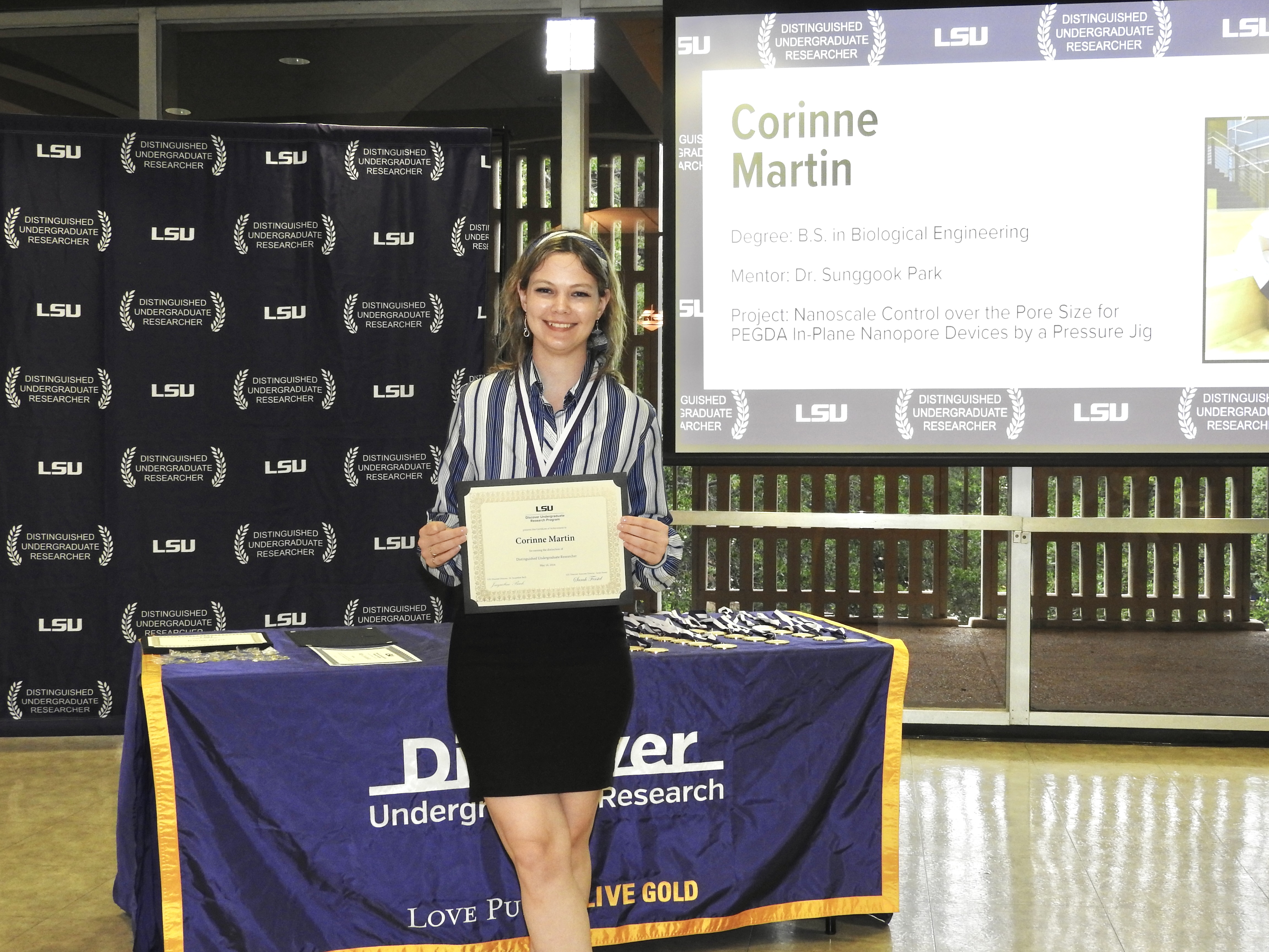 "Medium shot of Corinne Martin holding her Distinguished Undergraduate Researcher certificate in front of a screen displaying her work"